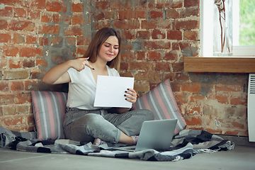 Image showing Young woman, businesswoman working or studying at home, looking on computer screen, monitor, holding white sheet, whiteboard. Attented, concentrated. Copyspace. Top view.