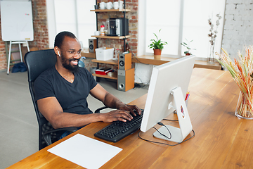 Image showing Young man, businessman working in office, looking on computer screen, monitor, with blank white sheet, whiteboard near him. Copyspace.