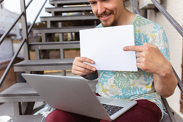 Image showing Young man, businessman working outdoors, looking on computer screen, monitor, holding blank white sheet, whiteboard. Copyspace.