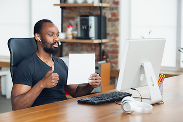 Image showing Young man, businessman working in office, looking on blank black computer screen, monitor, holding blank white sheet, whiteboard. Copyspace.