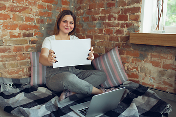 Image showing Young woman, businesswoman working or studying at home, looking on computer screen, monitor, holding white sheet, whiteboard. Attented, concentrated. Copyspace. Top view.