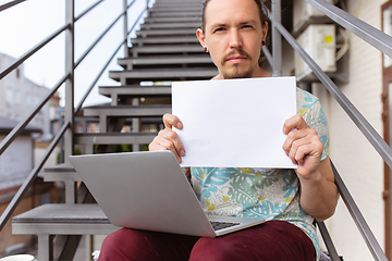 Image showing Young man, businessman working outdoors, looking on computer screen, monitor, holding blank white sheet, whiteboard. Copyspace.