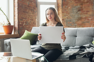 Image showing Young woman, businesswoman working or studying at home, looking on computer screen, monitor, holding white sheet, whiteboard. Attented, concentrated. Copyspace. Top view.