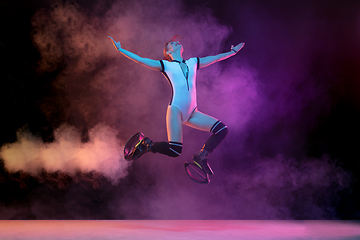 Image showing Beautiful redhead woman in a white sportswear jumping in a kangoo jumps shoes isolated on gradient studio background in neon lighted smoke
