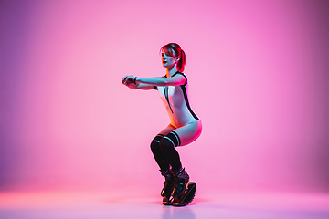 Image showing Beautiful redhead woman in a white sportswear jumping in a kangoo jumps shoes isolated on gradient studio background in neon light