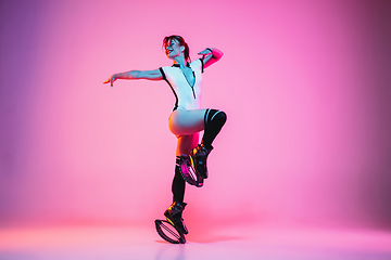 Image showing Beautiful redhead woman in a white sportswear jumping in a kangoo jumps shoes isolated on gradient studio background in neon light