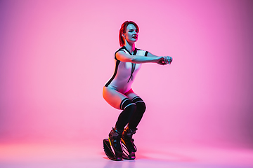 Image showing Beautiful redhead woman in a white sportswear jumping in a kangoo jumps shoes isolated on gradient studio background in neon light