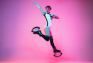 Image showing Beautiful redhead woman in a white sportswear jumping in a kangoo jumps shoes isolated on gradient studio background in neon light