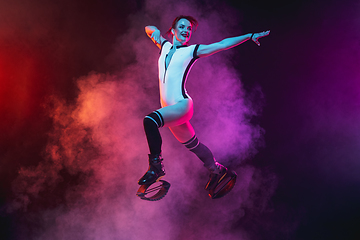 Image showing Beautiful redhead woman in a white sportswear jumping in a kangoo jumps shoes isolated on gradient studio background in neon lighted smoke