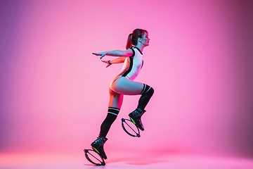 Image showing Beautiful redhead woman in a white sportswear jumping in a kangoo jumps shoes isolated on gradient studio background in neon light