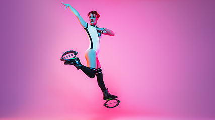 Image showing Beautiful redhead woman in a white sportswear jumping in a kangoo jumps shoes isolated on gradient studio background in neon light