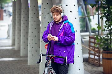 Image showing Handsome young man using mobile phone and headphones while standing near his bicycle beside him