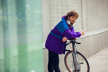 Image showing Handsome young man using mobile phone and headphones while standing near his bicycle beside him