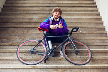 Image showing Handsome young man using mobile phone and headphones while sitting near his bicycle beside him