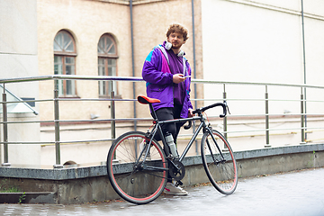 Image showing Handsome young man using mobile phone and headphones while standing near his bicycle beside him