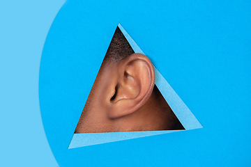 Image showing Ear of african-american man peeking throught triangle in blue background