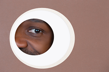 Image showing Eye of smiling african-american man peeking throught circle in brown background