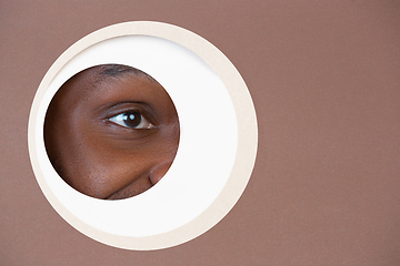 Image showing Eye of smiling african-american man peeking throught circle in brown background