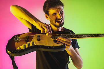 Image showing Young caucasian musician playing bass guitar in neon light on pink-green background