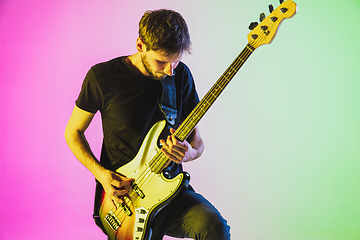 Image showing Young caucasian musician playing bass guitar in neon light on pink-green background