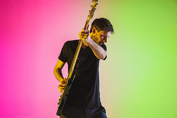 Image showing Young caucasian musician playing bass guitar in neon light on pink-green background