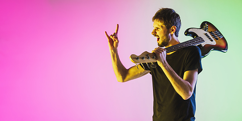 Image showing Young caucasian musician playing bass guitar in neon light on pink-green background