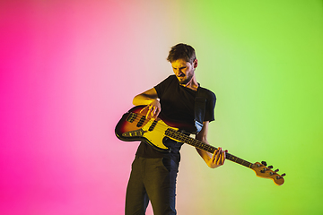 Image showing Young caucasian musician playing bass guitar in neon light on pink-green background