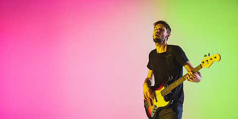 Image showing Young caucasian musician playing bass guitar in neon light on pink-green background