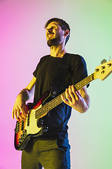 Image showing Young caucasian musician playing bass guitar in neon light on pink-green background