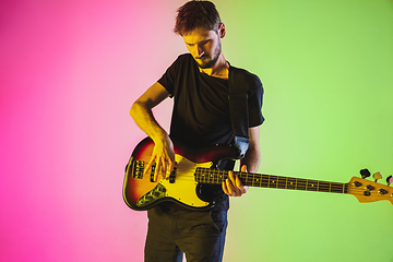 Image showing Young caucasian musician playing bass guitar in neon light on pink-green background