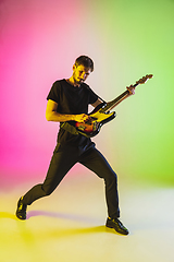 Image showing Young caucasian musician playing bass guitar in neon light on pink-green background