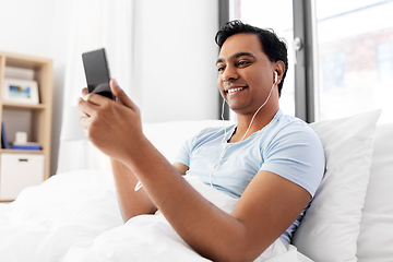 Image showing indian man in earphones with phone in bed at home