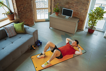 Image showing Young caucasian man training at home during quarantine of coronavirus outbreak, doing exercises of fitness, aerobic. Staying sportive during insulation.