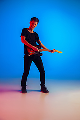 Image showing Young caucasian musician playing guitar in neon light on blue background, inspired