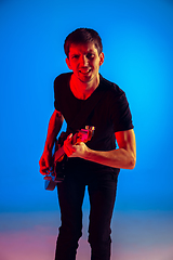Image showing Young caucasian musician playing guitar in neon light on blue background, inspired