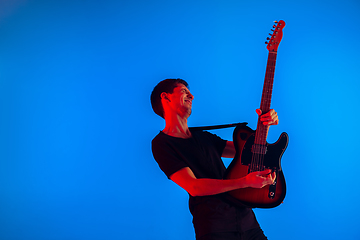 Image showing Young caucasian musician playing guitar in neon light on blue background, inspired