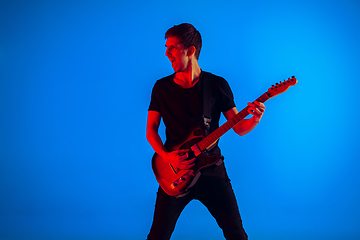 Image showing Young caucasian musician playing guitar in neon light on blue background, inspired