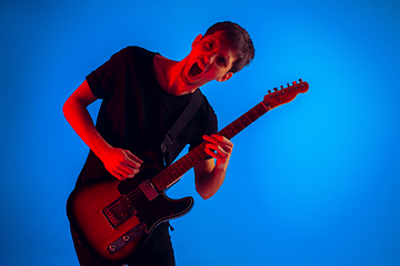 Image showing Young caucasian musician playing guitar in neon light on blue background, inspired