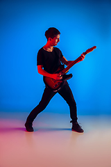 Image showing Young caucasian musician playing guitar in neon light on blue background, inspired