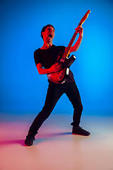 Image showing Young caucasian musician playing guitar in neon light on blue background, inspired