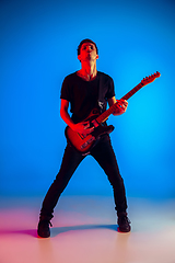 Image showing Young caucasian musician playing guitar in neon light on blue background, inspired