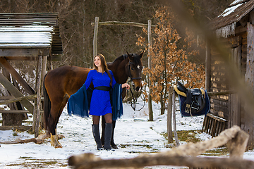 Image showing A beautiful girl in a blue dress stands with a horse against the background of old wooden buildings in a winter forest