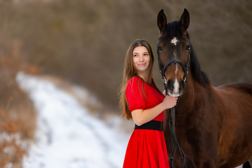 Image showing Portrait of a beautiful girl in a red dress standing with a horse on the background of a winter road