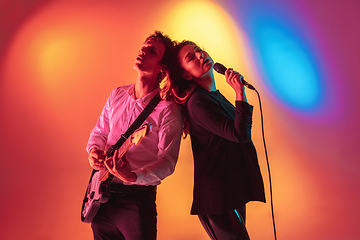 Image showing Young caucasian musicians, female singer and guitarist performing on gradient background in neon light. Concept of music, hobby, festival