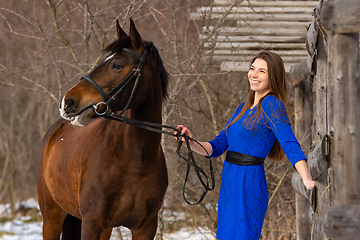 Image showing A happy girl holds a horse by the bridle and smiles, against the backdrop of a winter forest