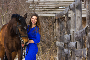 Image showing A beautiful young girl in a blue dress hugs a horse against the background of an old fence and a winter forest