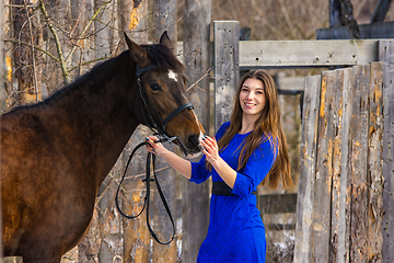 Image showing Portrait of a beautiful girl in a blue dress with a horse against the background of an old wooden fence