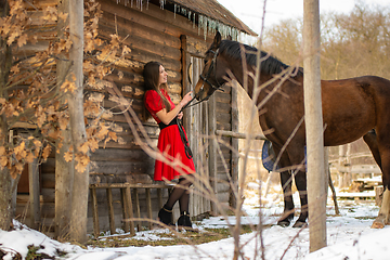 Image showing A girl in a red dress stands near an old wooden house, a horse stands next to her.