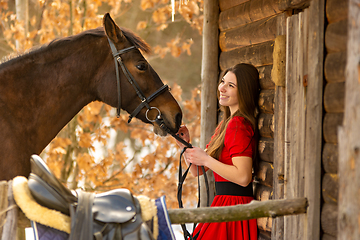 Image showing Portrait of a beautiful girl in a red dress, a girl holding a horse by the bridle, a beautiful background of a wooden wall and forest