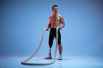 Image showing Athlete with disabilities or amputee isolated on blue studio background. Professional male sportsman with leg prosthesis training with weights in neon